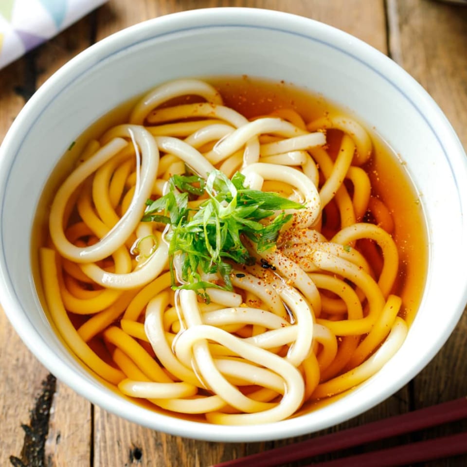 A close-up of a bowl of udon noodles in broth, garnished with green onions and spices.
