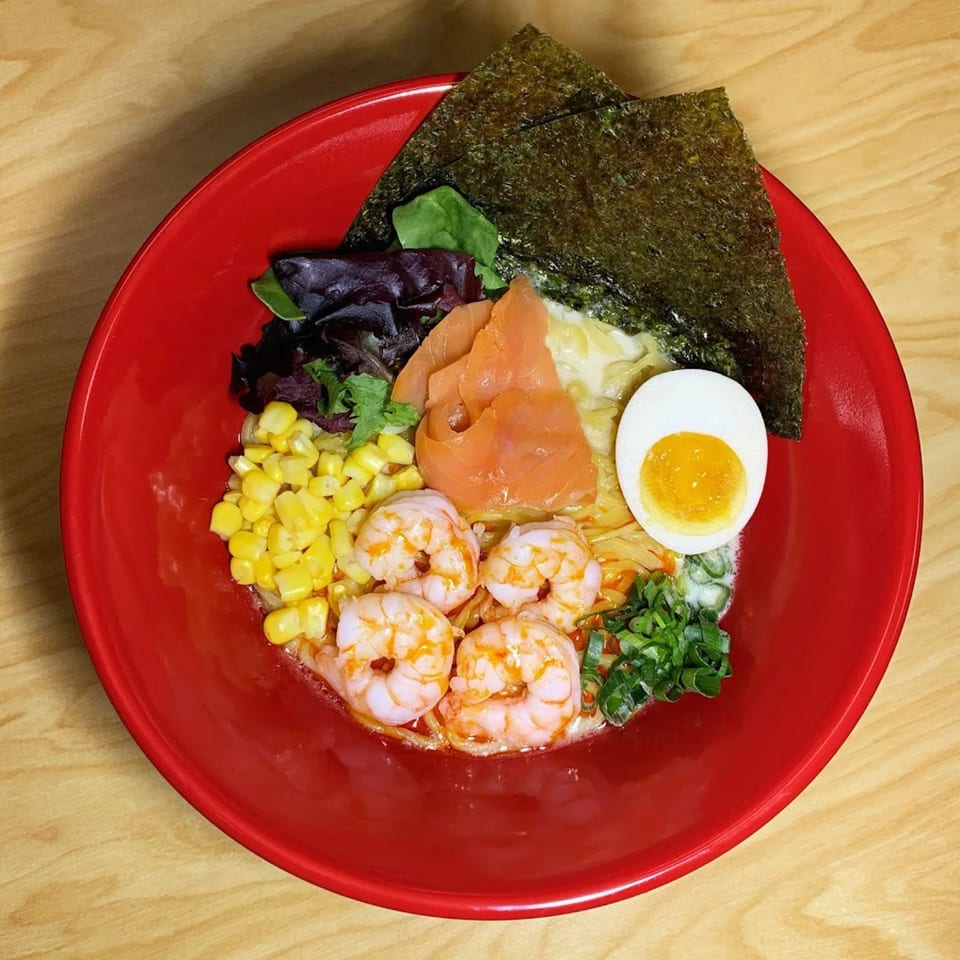 Aroma Ramen's ramen bowl with shrimp, salmon, corn, nori, greens, and a soft-boiled egg in a red bowl.
